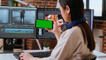Asian female editor working from home for freelance work with green screen, using smartphone and dual monitors for mixing software. Creative workspace equipped with tech tools. Camera B. photo