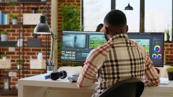 Male editor works from modern home office in the living room, multitasking with a dual monitor setup and editing software. Finishing post production tasks for media production. Camera B. photo