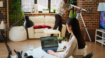 Software developer types programming code in a cozy home office setup. Asian woman coding for cloud computing on her laptop, enhances productivity for software development. Camera A. photo