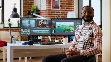African american male filmmaker works with creative editing software, mixing and color grading clips. Living room workspace features post production tools and dual monitors. Camera B. photo