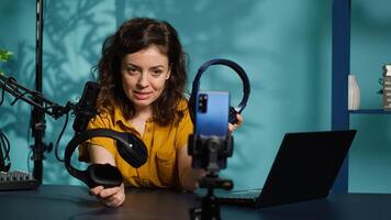 Audio engineer recording for internet channel in dimly lit home studio, presenting pairs of gaming headphones to audience. Woman showing viewers music listening device, camera A photo