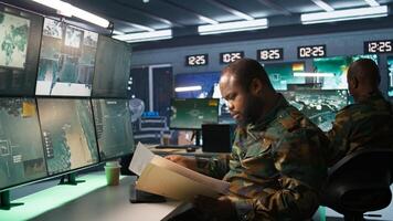 Military soldier in mission control center overseeing national safety operations, reading folder files. African american army specialist checking data in monitoring room command center, camera B photo