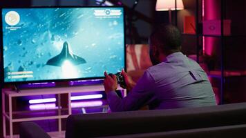 Gamer using controller and headphones to play first person shooter videogame on large smart TV. Man in home theatre using high tech gaming console gamepad to defeat foes, camera A photo