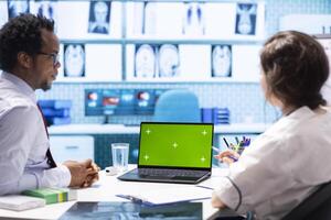 General practitioner and patient looking at green screen on device, recommending pills and drugs during check up visit. Diverse people discussing about diagnostics and remedy. photo