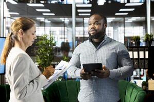 Employee presenting documents to manager holding tablet, showing him researching key data needed for finding business insights. Company executives analyzing data on device and paperwork photo