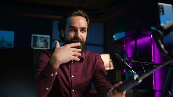 Happy influencer in home studio greeting audience, recording live broadcast using smartphone on selfie stick, close up. Man filming q and a session with mobile phone, answering questions, camera B photo