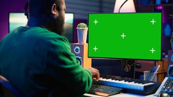 Musician operates on midi controller piano keys next to mockup screen, producing new songs with musical instruments and audio gear in home studio. Artist works with green screen. Camera A. photo