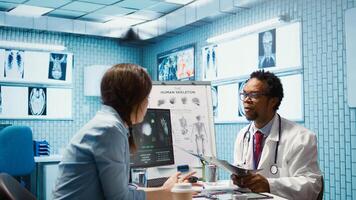 Female patient listening to a diagnostic report from doctor based on MRI scan results and x ray imaging, pointing at computer in medical cabinet. African american expert gives assistance. Camera B. photo