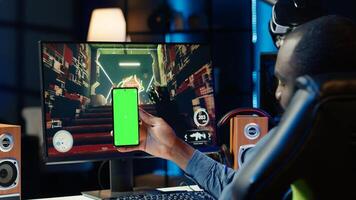 Man texting friends on green screen phone while playing first person shooter videogame at home. Gamer looking at mockup smartphone display, enjoying online discussion with mates photo