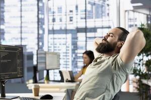 Fatigued admin fixing bad posture while managing cloud computing platforms to train machine learning models. Tired man stretching, trying to relax while monitoring and maintaining company systems photo