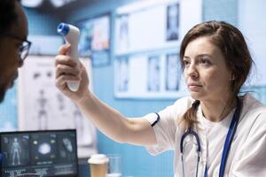 Healthcare specialist using thermometer to check fever measurements, taking temperature levels at a medical consultation. Young medic using digital instrument for diagnostic reports. photo