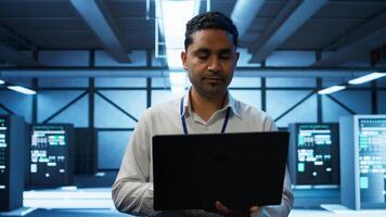 System administrator in data center using notebook to design, implement and manage gear, ensuring optimal performance and reliability. Indian man in server room coding on laptop, camera B photo