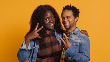 Cute life partners posing in studio and doing peace sign in studio, showing the victory symbol against yellow background. African american spouses doing kawaii gesture. Camera A. photo