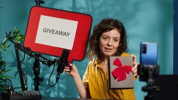 Smiling woman holding empty copy space sign, excited to do giveaway for subscribers. Media star happy to offer packed gift with ribbon to viewers, holding cardboard bubble speech cutout, camera A photo