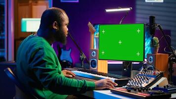 Songwriter operates on synthesizer piano keys next to isolated display, composing new music with instruments and stereo tools in home studio. Artist producer works with green screen. Camera B. photo
