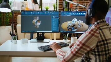 African american industrial designer works on CAD software and listens to music, multitasking at home. Engineer specialist uses digital tools and 3D modeling for industrial projects. Camera A. photo