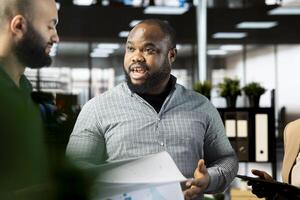 African american employee in company office checking progress on customer commissioned project. Man collaborating with colleague, comparing key data statistics after business audit photo