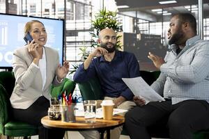 Happy woman talking on phone next to coworkers looking over revenue figures on documents in office. Cheerful worker in telephone call next to colleagues comparing company statistical numbers photo