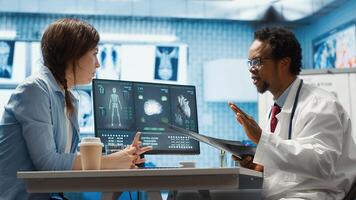 Female patient listening to a diagnostic report from doctor based on MRI scan results and x ray imaging, pointing at computer in medical cabinet. African american expert gives assistance. Camera A. photo