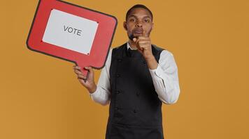 Catering employee showing a message to vote on red speech bubble, presenting the cardboard icon to do a commercial for politics. Waiter in uniform works on customer service. Camera B. photo