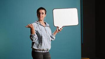 Female model with a groovy style holding a speech bubble in studio, creating an ad commercial with an empty billboard icon. Cool edgy woman presenting a sign for advertisement. Camera A. photo