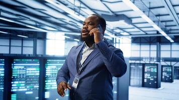 IT support worker in data center talking in phone call, doing yearly checkup. On field engineer chatting with manager in server hub during telephone call, monitoring servers infrastructure, camera B photo