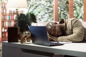 Small business owner feels tired and takes a nap while doing remote work, sleeping on his home desk. Stressed overworked freelancer falling asleep at the office setup, financial operations. photo
