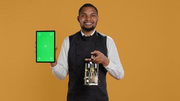 Professional waiter with apron presenting tablet with isolated mockup, presenting the copy space while he carries the cruet set. Skilled catering staff works in customer service at diner. Camera B. photo