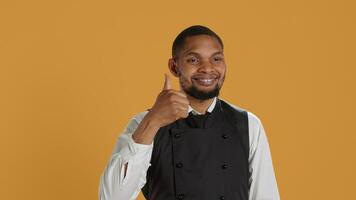 Waiter smiling and showing a thumbs up symbol against yellow background, presenting his satisfaction and approval with a positive like sign. Cheerful staff gives okay gesture. Camera A. photo