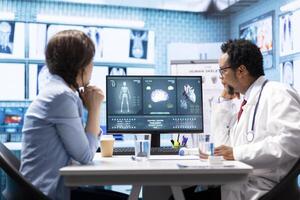 Physician reviewing MRI scan and x ray test results with a patient in medical office, explaining the diagnostics. Doctor pointing at analysis to discuss about the illness, medical technology. photo