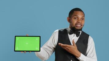 Doorkeeper bellboy poses with green screen on tablet at the hotel reception, working in hospitality industry and greeting customers. Lodging resort personnel shows chroma key. Camera A. photo