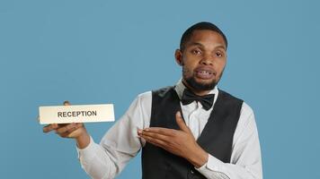 Hotel concierge staff showing the reception indicator at front desk, greeting guests at five star holiday resort in studio. Skilled professional bellboy holds sign against blue background. Camera A. photo