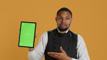 Restaurant waiter presenting a green screen layout on tablet against yellow background, wearing professional service oriented attire on duty. Butler serving clients, professionalism. Camera A. photo