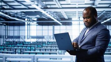 African american IT operator doing maintenance using laptop to prolong data center equipment life span. IT professional in server hub workspace monitoring server mainframes using notebook, camera B photo