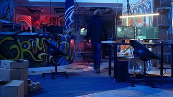 Hooded hacker arrives in neon lit underground shelter filled with graffiti artwork. Cybercriminal enters colorful painted basement, starting work on script that can crash businesses servers, camera A photo