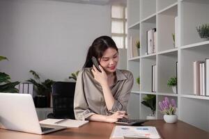 Young Asian business woman sits on the phone in an online business meeting using a laptop in a modern home office decorated with shady green plants. photo