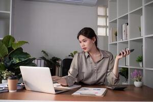 Young Asian business woman sits on the phone in an online business meeting using a laptop in a modern home office decorated with shady green plants. photo