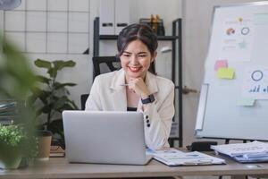 Asian businesswoman or accountant happily sitting and working with laptop on finance and business administration. On the desk in the office decorated with green plants. photo
