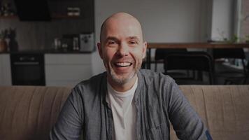 A man sits on a sofa in a cozy kitchen, wearing a white T-shirt, gray shirt, and jeans, smiling warmly, creating a friendly atmosphere. Background includes shelves video