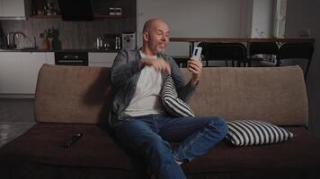 A middle-aged man sits in his living room, having a call using his smartphone. The kitchen and home environment are visible in the background. He is in a good mood, enjoying the conversation. video