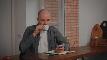 A middle-aged man relaxes in his kitchen, enjoying a cup of espresso coffee. He is scrolling through content on his smartphone, browsing social media. video