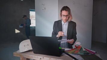 A woman interior designer is working on creating a design project at a construction site. The specialist uses a laptop, drawings and palettes to find the style of the room. video