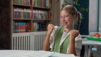 Excited happy lucky little girl Caucasian schoolgirl kid school child student pupil learner looking at camera gesture clenching fists celebrating yes win successful pass test exam in library classroom video
