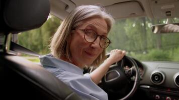 Back view happy smiling Caucasian elderly mature senior woman in glasses taxi car driver female automobile owner rental service client turning head looking camera smile inside vehicle transportation video