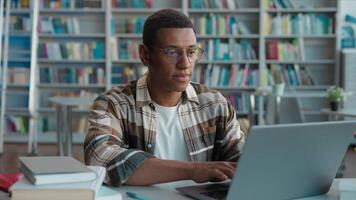 African American man guy male typing browsing laptop online computer high school library university student studying learning e-learning looking at camera smiling internet networking texting college video