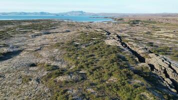 Icelandic Plate Fault. A large, rocky field with a body of water in the background. The field is covered in grass and plants, and the water is calm and still video
