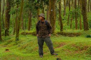 An adventurer in a flannel shirt, bucket hat and backpack poses against a backdrop of dense pine trees. Asian man in adventure clothes in dense and green pine forest area. photo