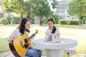 Asian kid girl and friend learning online guitar lesson in the park, Friends playing guitar on picnic in park photo