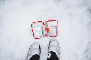 An open first aid kit with medicines lies on the snow at the feet of a man, top view of pills in a red first aid kit. photo