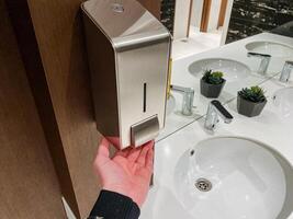 A person using a soap dispenser in a bathroom with hardwood flooring and wood stain on the cabinetry of a building photo